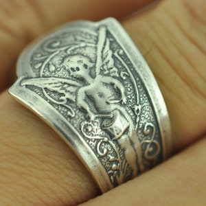 May include: A silver ring with an ornate design featuring a cherub with wings. The cherub is holding a heart in its hands.