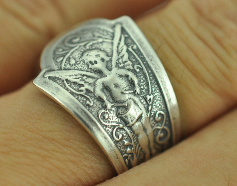 May include: A silver ring with an ornate design featuring a cherub with wings. The cherub is holding a heart in its hands.