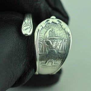 May include: A silver-toned spoon ring featuring an embossed design of the Libra zodiac sign. The center displays a hand holding a scale above a sunburst, with the word LIBRA inscribed at the top and the Libra symbol engraved at the bottom.