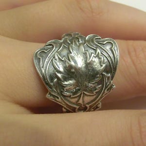 May include: A silver-toned ring featuring an embossed maple leaf design at the centre. The metal surface has a darkened, oxidised finish within the recessed areas, highlighting the intricate, swirling scrollwork patterns surrounding the leaf.