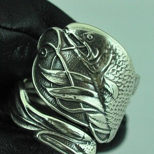 May include: A silver-toned metal ring featuring a detailed relief design of a fish swimming among aquatic plants. The band of the ring is textured with a pattern resembling flowing water or reeds, wrapping around the finger with a metallic finish.