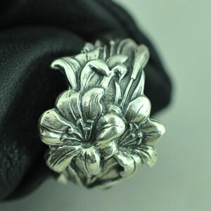 Puede incluir: Un anillo de plata con un diseño floral tridimensional detallado que presenta múltiples pétalos de lirio superpuestos. El metal tiene un acabado pulido con áreas hundidas oscurecidas que resaltan las curvas y texturas de los pétalos.