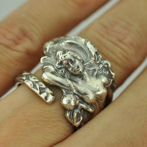 May include: A silver-toned metal ring featuring an intricate relief design of a figure with flowing hair and a smaller cherub-like form. The band has a textured, leaf-like pattern and an open-ended design that wraps around the finger.