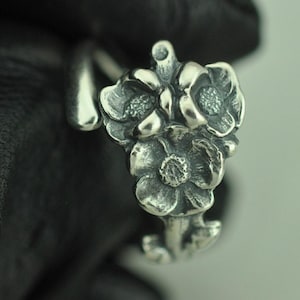Könnte beinhalten: Ein silberfarbener Ring mit einem Cluster aus dreidimensionalen Blumen mit texturierten Mitten und glatten Blütenblättern. Die Ringschiene ist wie ein schlanker Stängel geformt, der sich um den Finger legt und ein poliertes Metallfinish aufweist.