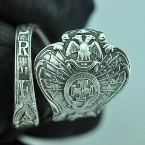 Può includere: Un anello in argento con una fascia larga che presenta il testo in rilievo PROOF OF FAITH e una grande parte frontale ornata che mostra un emblema con aquila a due teste sopra un simbolo a croce, circondato da raggi decorativi e dettagli.