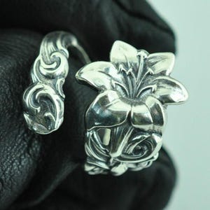 May include: A silver-toned open ring featuring a detailed lily flower motif on one end and a swirling, scroll-like pattern on the other. The metal surface has a polished finish with darkened recessed areas that highlight the intricate floral design.