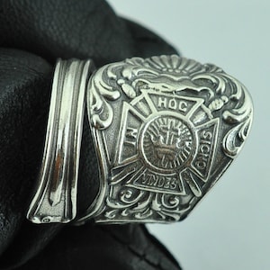 Könnte beinhalten: Silberring mit einem detaillierten Design, das ein Kreuz mit den Worten "HOC SIGNO VINCES" und eine Krone zeigt. Der Ring ist vor einem schwarzen Lederhintergrund platziert.