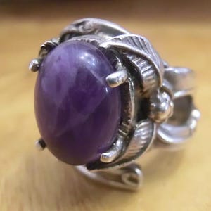 May include: A silver ring featuring a large, oval-shaped, deep purple gemstone. The ring has an ornate design with leaf and floral details surrounding the stone. The ring is set against a warm, wooden background.
