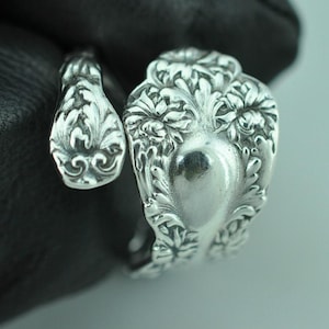 May include: A silver-toned metal ring crafted from a repurposed spoon, featuring an open-ended design. The surface displays intricate, raised floral and leaf-like embossed patterns that cover the entire exterior of the band and the curved ends.