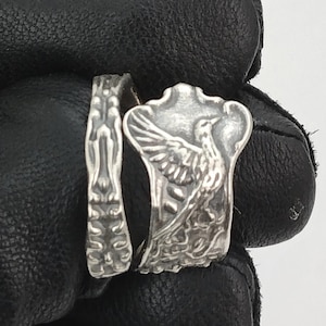 May include: Two silver rings, one with a detailed design of a bird with outstretched wings and the other with a floral pattern.