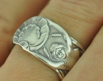 Puede incluir: Un anillo de tono plateado con un diseño en relieve de una figura con cabello fluido rodeada de elementos florales estilizados y espadañas. La superficie metálica tiene un acabado mate con áreas hundidas oscurecidas.