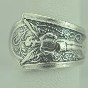 May include: Silver ring with an angel design. The angel is depicted with wings and a halo, and is surrounded by intricate floral details.