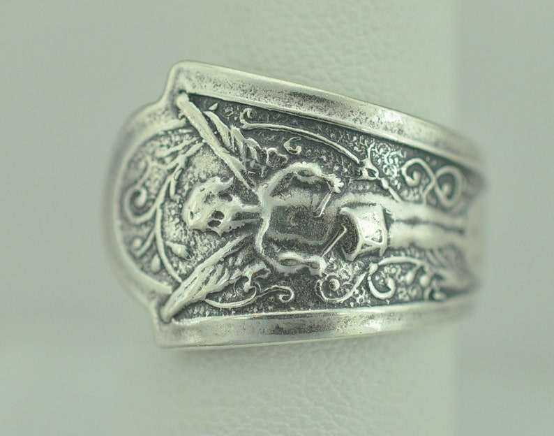 May include: Silver ring with an angel design. The angel is depicted with wings and a halo, and is surrounded by intricate floral details.