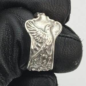 May include: A silver ring with a detailed design of a bird with outstretched wings. The bird is perched on a branch with leaves.