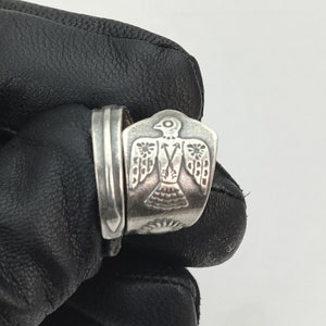 Op de afbeelding: Een zilveren ring met een gedetailleerd ontwerp van een vogel met uitgespreide vleugels. De vogel heeft een kleine cirkel voor een oog en een kleine pijl in zijn borst.