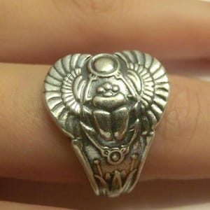 May include: A silver-toned ring featuring a detailed relief of a scarab beetle with outstretched wings. The design includes a circular sun disk above the beetle and stylized lotus flower motifs at the base of the band, crafted in a metallic finish.