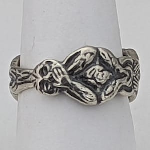 May include: A silver ring with a detailed design of intertwined figures. The ring is made of metal and has a smooth, polished finish.