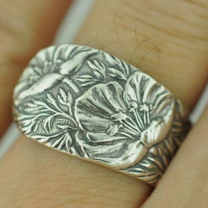 May include: A wide silver ring featuring a detailed, raised relief pattern of blooming flowers and foliage. The metal has a polished finish with darkened recessed areas that highlight the intricate botanical design across the entire band surface.