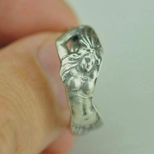 May include: A silver-toned metal ring featuring a sculpted relief of a figure with long flowing hair, a defined torso, and a textured lower section. The design wraps around the band, showcasing intricate details in the metalwork and oxidized finish.