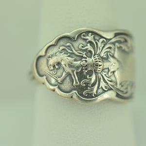 May include: A silver ring with a detailed design of a lion holding a sword and a crown. The ring is made from a repurposed spoon.