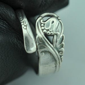 May include: A silver-toned spoon ring with an ornate floral design. The ring features a detailed, openwork floral motif on the face and a curved band. The spoon bowl is visible on the side, adding to the unique design.