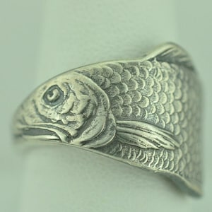 以下が含まれることがあります： 魚の頭と鱗の精巧なエンボス加工が施された銀色の金属製リング。表面の凹んだ部分には暗い酸化仕上げが施されており、魚の目、エラ、ヒレ、重なり合う鱗の質感を強調しています。