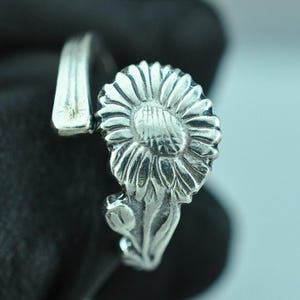 May include: A silver-toned ring crafted from a repurposed spoon, featuring a prominent embossed sunflower design on the face. The band is formed from the handle of the spoon, showing a smooth, curved metal surface with a polished finish.