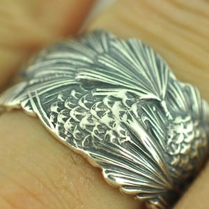 May include: A wide silver ring featuring a detailed, raised relief pattern of overlapping pine needles and pine cones. The metal has a darkened, oxidized finish in the recessed areas to highlight the intricate texture of the botanical design.