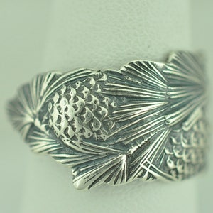 May include: A silver ring with a detailed design of pine needles and pine cones.