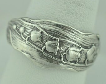 Peut inclure: Une bague en métal argenté présentant un motif en relief de fleurs et de feuilles de muguet. La surface possède une finition oxydée qui souligne les détails botaniques complexes du motif floral qui fait le tour de l'anneau.