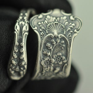 May include: A silver ring crafted from a repurposed spoon handle, featuring an intricate embossed floral pattern with a central shell motif and scrolling details. The metal has a darkened, oxidized finish that highlights the relief texture.