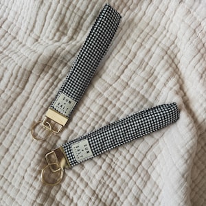 May include: Two fabric wristlet keychains featuring a black and white gingham check pattern. Each keychain has a gold-toned metal clasp and a circular ring. A small white fabric label is sewn onto each strap with the text SUN + SALT DESIGN.