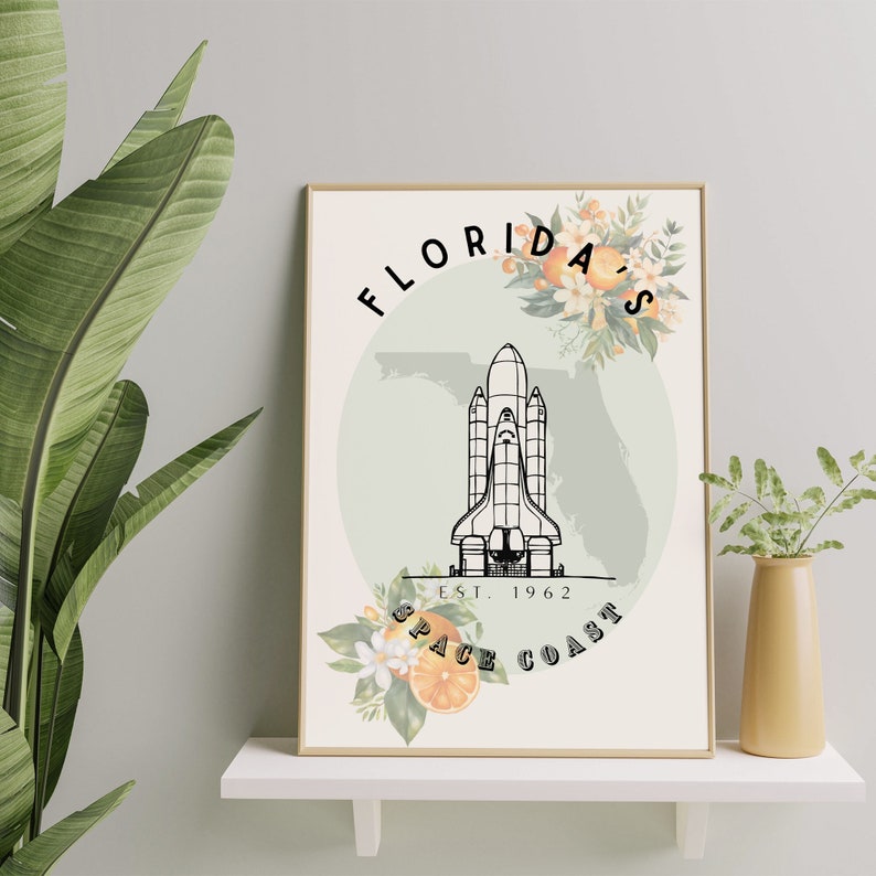 May include: A framed art print featuring a black and white illustration of a space shuttle, with the text "Florida's Space Coast Est. 1962." The design includes orange blossoms and a light green outline of Florida.