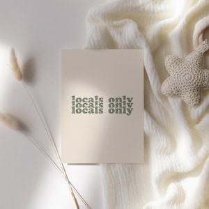 May include: A cream-colored print with the words "locals only" in a repeating design, in a muted green font. The print is on a white surface with a white, textured fabric and a star-shaped baby toy.