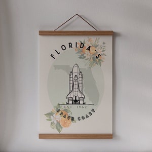May include: A poster with a wooden frame and a hanging string. The poster features a black and white illustration of a space shuttle, the text "Florida's Space Coast Est. 1962", and floral accents with oranges.