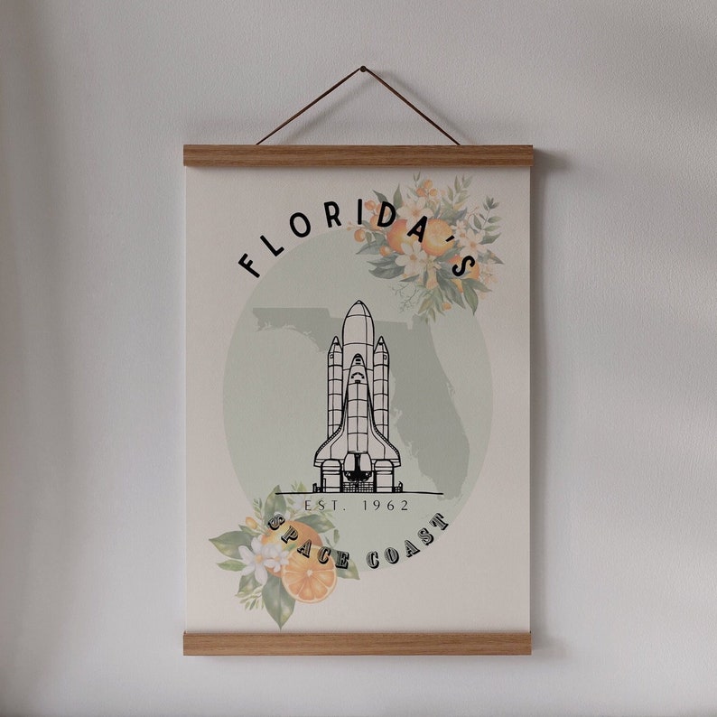 May include: A poster with a wooden frame and a hanging string. The poster features a black and white illustration of a space shuttle, the text "Florida's Space Coast Est. 1962", and floral accents with oranges.