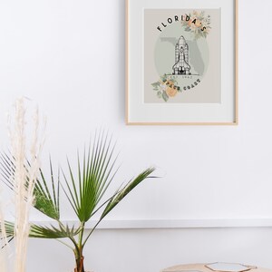 May include: Framed art print featuring a black and white illustration of a space shuttle over a map of Florida. The words "Florida Space Coast" are displayed with floral accents. The print is in a light wood frame.