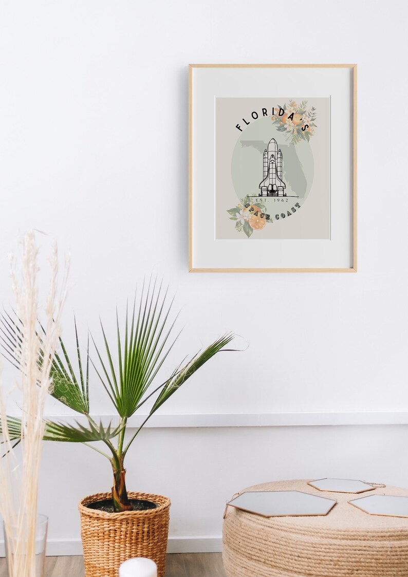 May include: Framed art print featuring a black and white illustration of a space shuttle over a map of Florida. The words "Florida Space Coast" are displayed with floral accents. The print is in a light wood frame.