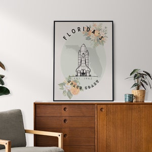 May include: Black framed art print featuring a space shuttle illustration with the text "Florida's Space Coast Est. 1962". The design includes floral and orange slice accents, set against a light green backdrop.