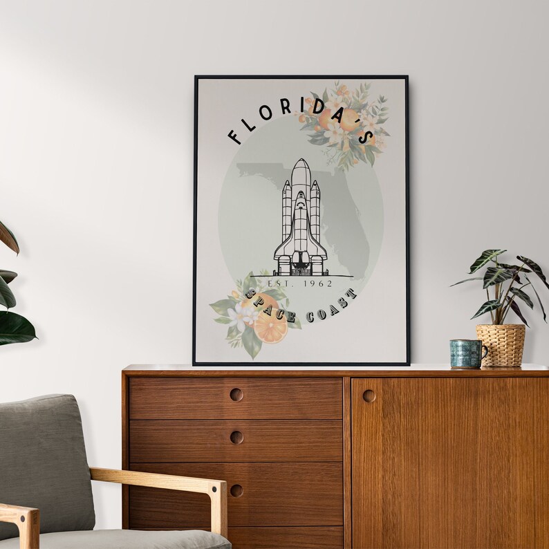 May include: Black framed art print featuring a space shuttle illustration with the text "Florida's Space Coast Est. 1962". The design includes floral and orange slice accents, set against a light green backdrop.