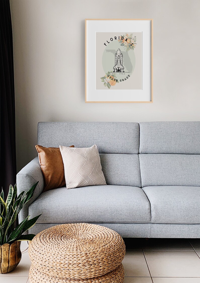 May include: A framed print featuring a Florida Space Coast design. The artwork includes a stylized illustration of a space shuttle, floral accents, and the text "Florida Space Coast." The print is displayed above a gray sofa with decorative pillows.