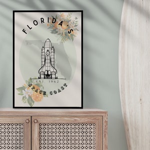 May include: Black framed art print featuring a space shuttle illustration with the text "Florida's Space Coast" and the year "Est. 1962". The design includes floral and orange accents, set against a light green background. The print is displayed on a wooden cabinet.