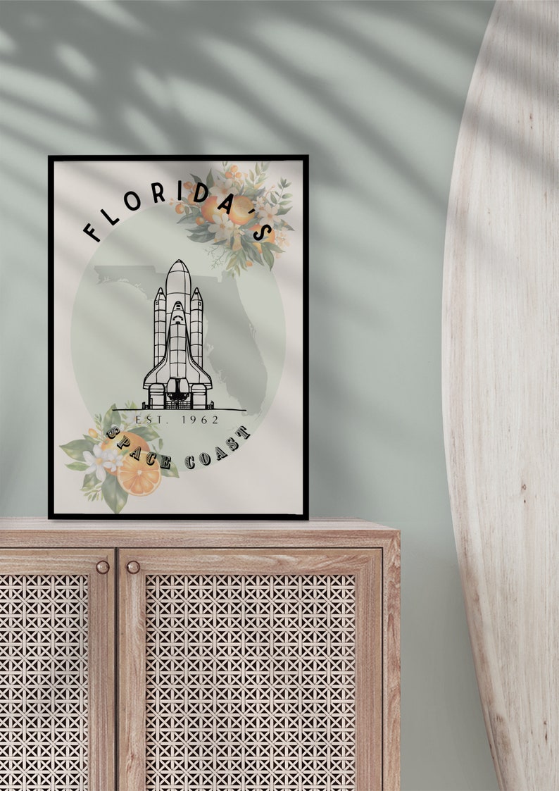 May include: Black framed art print featuring a space shuttle illustration with the text "Florida's Space Coast" and the year "Est. 1962". The design includes floral and orange accents, set against a light green background. The print is displayed on a wooden cabinet.