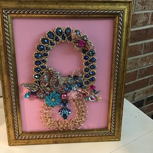 Op de afbeelding: Een ingelijst kunstwerk met een roze achtergrond en een goudkleurige lijst. Het kunstwerk toont een decoratief ontwerp gemaakt van blauwe en roze juwelen, een gouden ketting en bloemaccenten. Het ontwerp heeft de vorm van een acht.