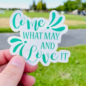 May include: White sticker with the text "Come! What may and love it" in teal green lettering. The sticker is held in a hand against a green grassy background.