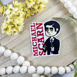 May include: A sticker featuring a cartoon illustration of Michael Scarn from the TV show The Office. The sticker is white with red text that says "MICHAEL SCARN". The cartoon character is wearing a blue suit and tie and holding two guns.