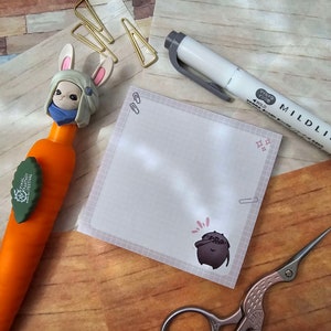 May include: A white and gray marker pen with the text "Mildliner" and "Zebra" on the side. The marker is on a wooden surface with a white and gray notepad with a small cartoon character on it. There are also gold paper clips and a pair of silver scissors on the surface.