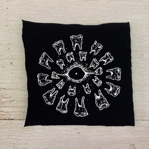 May include: Black fabric patch with a white embroidered design of a circle of teeth surrounding a single eye in the centre.
