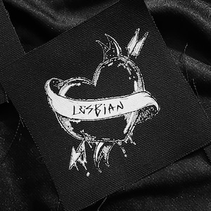 Könnte beinhalten: Schwarz-weiß gestickter Patch mit einem von einem Pfeil durchbohrten Herzen und einem Banner mit der Aufschrift "LESBIAN".