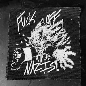 Pode incluir: Patch preto e branco com uma criatura semelhante a um lobo mordendo um braço. O texto "FUCK OFF NAZIS!" está escrito em negrito. O patch tem uma aparência áspera.
